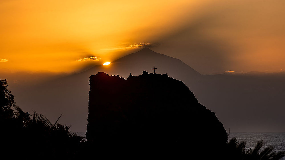 Blick von einer Klippe auf den Hafen von Puerto de las Nieves auf Gran Canaria. Im Vordergrund ragen vulkanische Felsen mit grüner Vegetation ins Bild, unten liegt der Hafen mit einem Fährschiff und weißen Häusern. Dahinter erstreckt sich die Küste bis zum Horizont, umgeben vom blauen Atlantik im warmen Abendlicht.